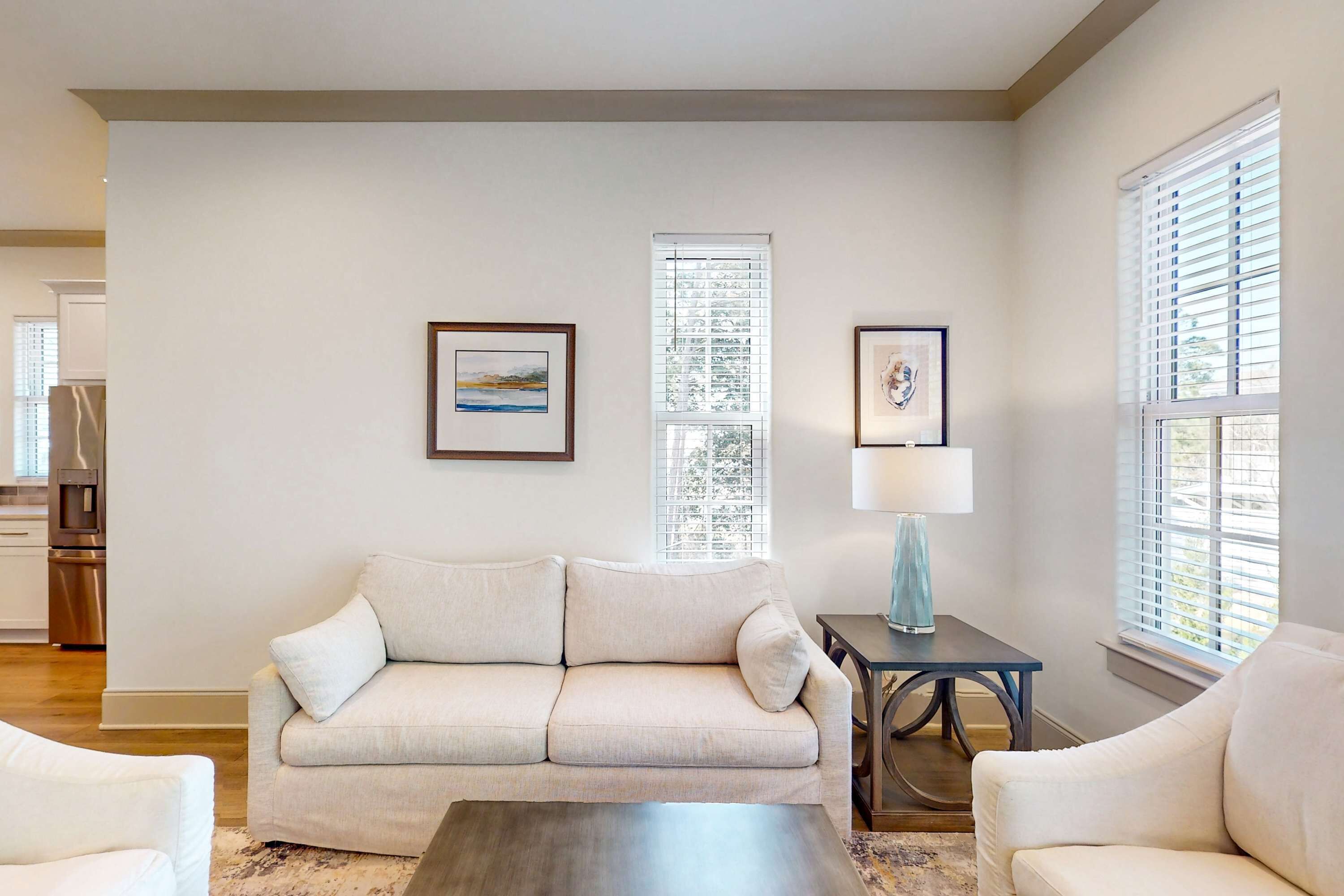 A living room with a beige sofa, a coffee table, and two side tables with lamps. There are framed pictures on the wall and a window with blinds.