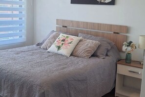 Un dormitorio con cama, mesita de noche, un jarrón con flores y una ventana con persianas.