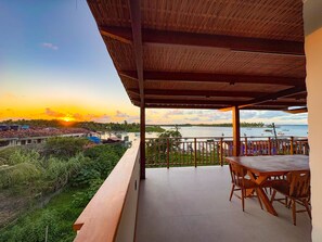 A balcony with a view of a sunset over a body of water, surrounded by greenery and buildings. - Pousada Feliz Boipeba (Cairu)