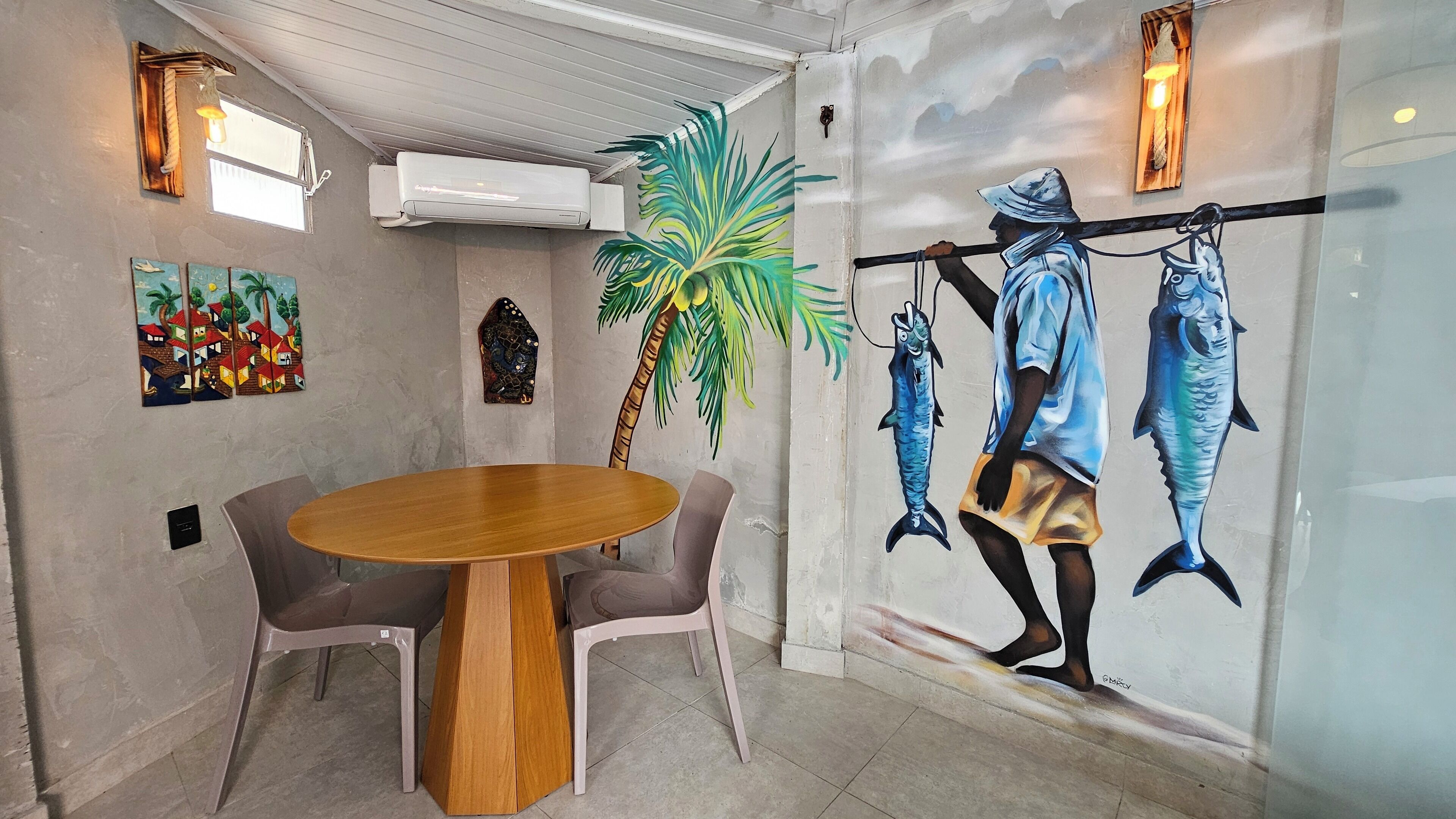 A dining area with a round wooden table and two chairs, a mural of a fisherman, and a palm tree.