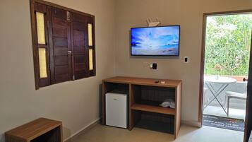 Habitación compacta con escritorio de madera, refrigerador blanco pequeño, televisor montado en la pared y puerta que da a un espacio exterior con vegetación.