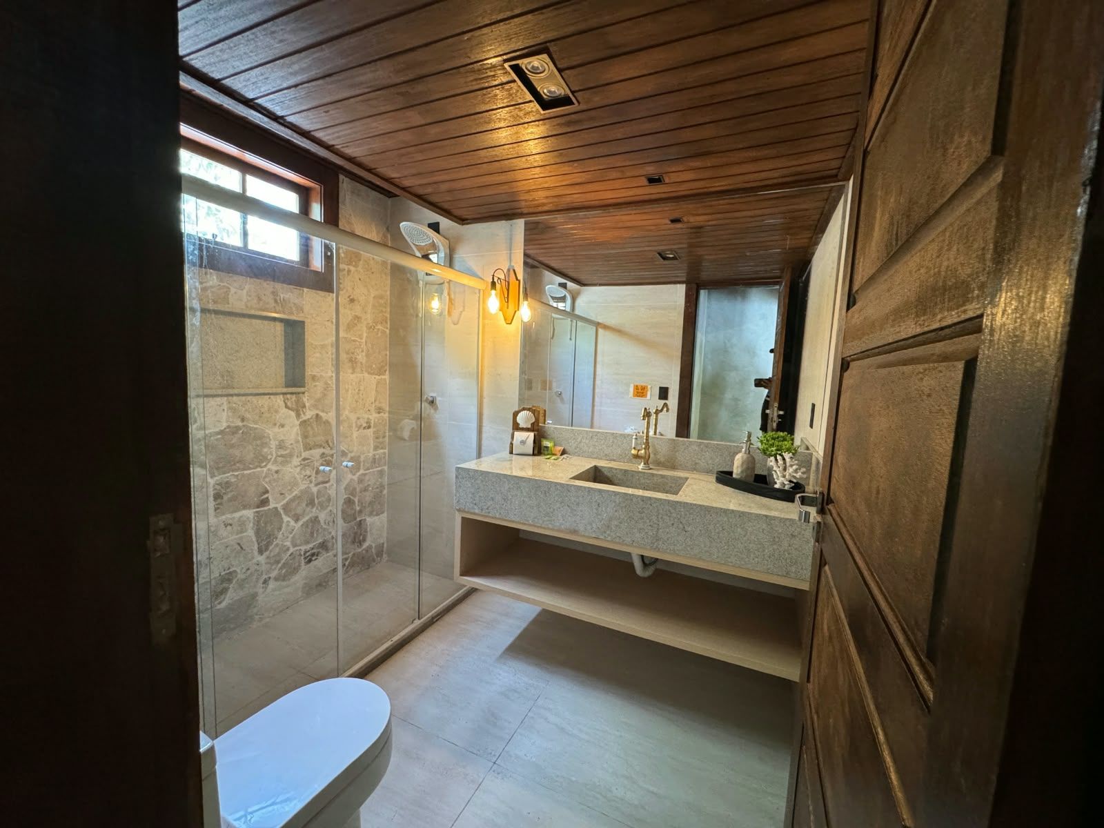 Une salle de bain avec une douche à l’italienne en pierre, un plafond en bois, un grand miroir et des toilettes blanches.