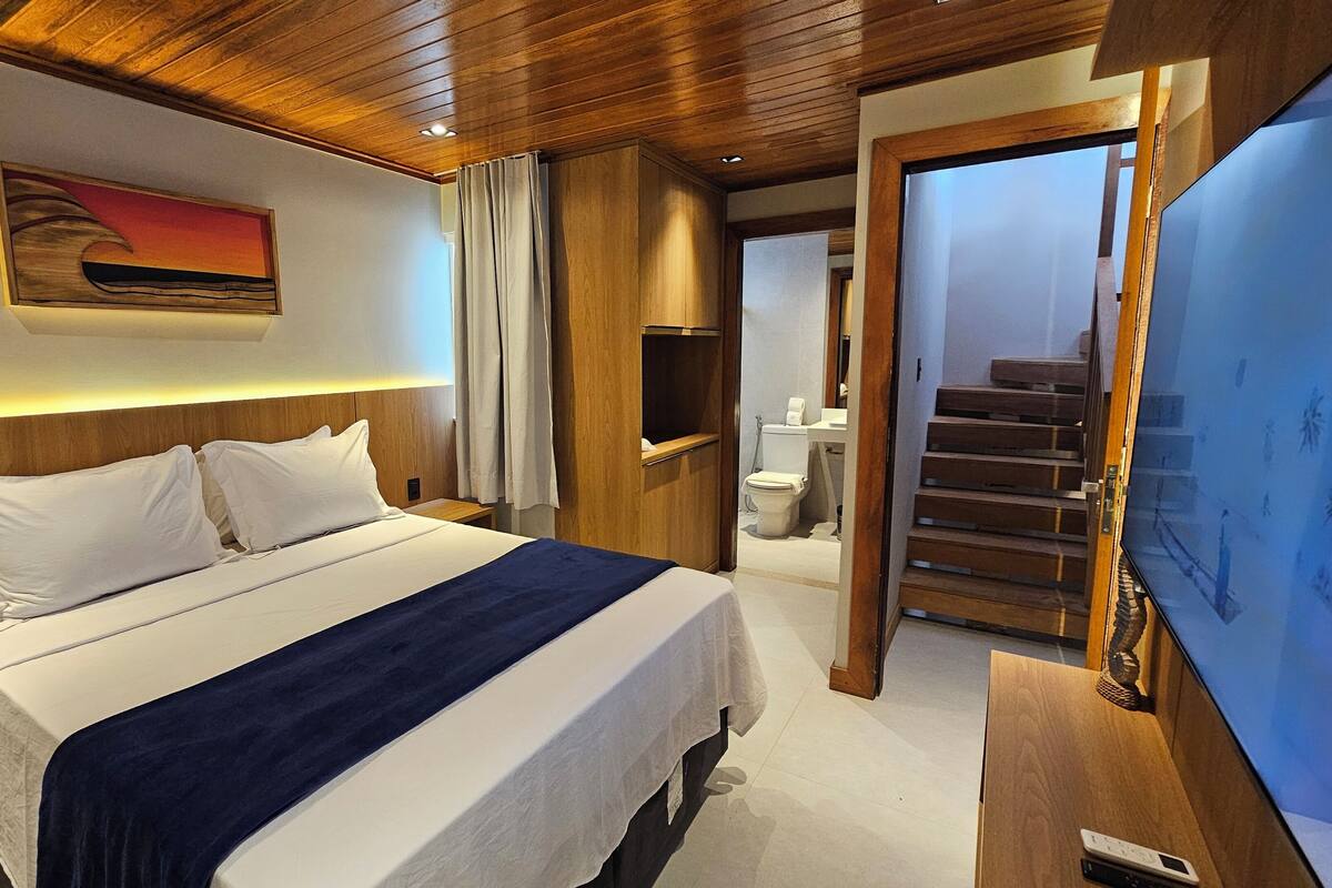 A hotel room with a bed, a TV, a wooden headboard, a painting, and a bathroom visible through an open door.