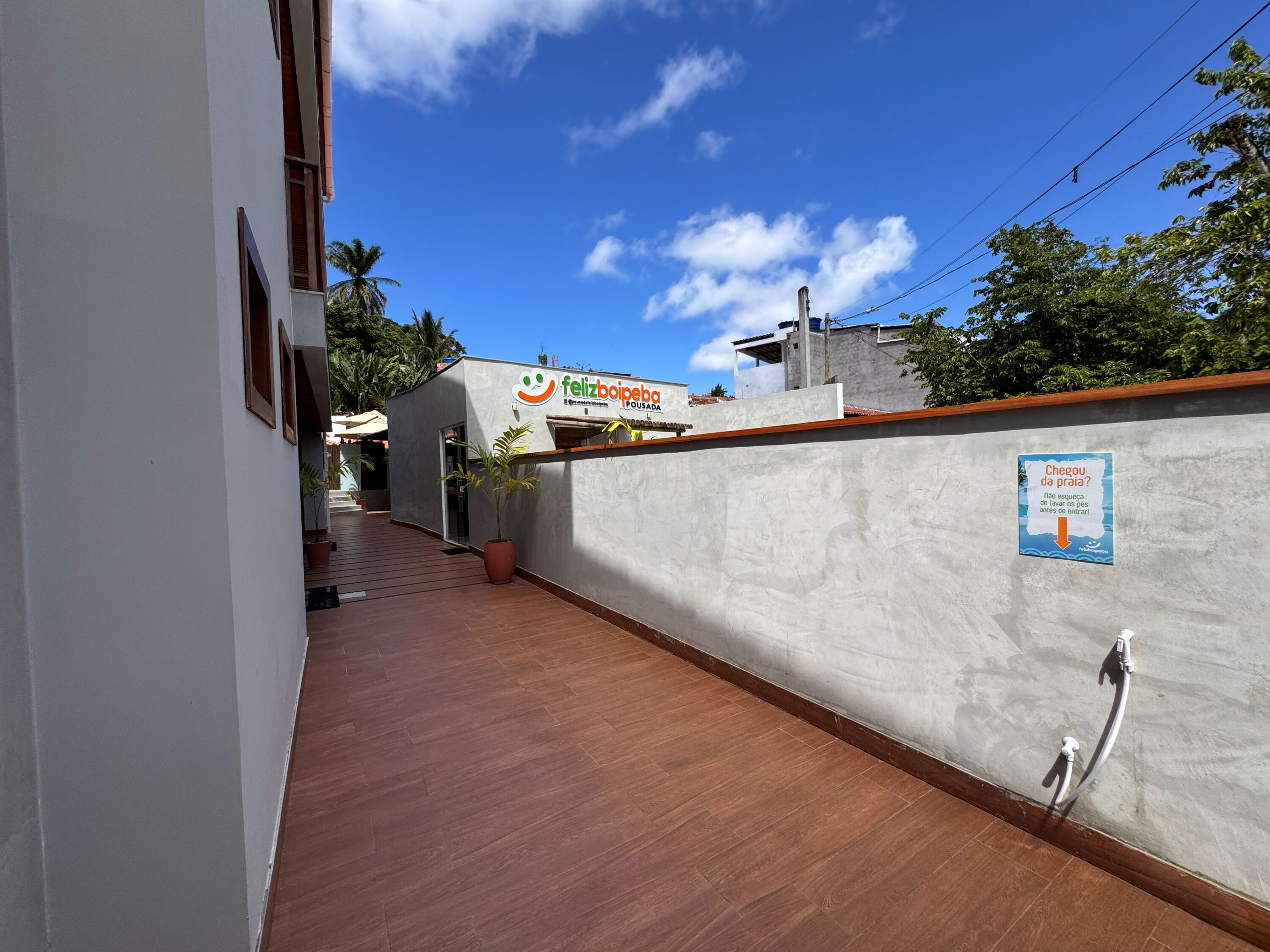 Um corredor estreito com piso de madeira, parede branca e uma placa com o nome 'felizboipeba' em um prédio ao fundo.