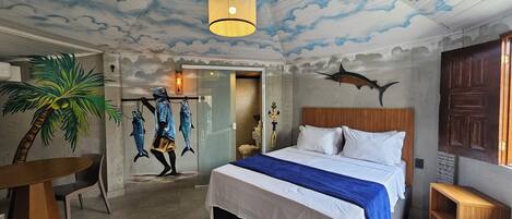 Un dormitorio con cama, cabecera de madera, mesita de noche, mesa, silla, lámpara de pared, techo con diseño de nubes, decoración de pez y un cuadro de palmera.