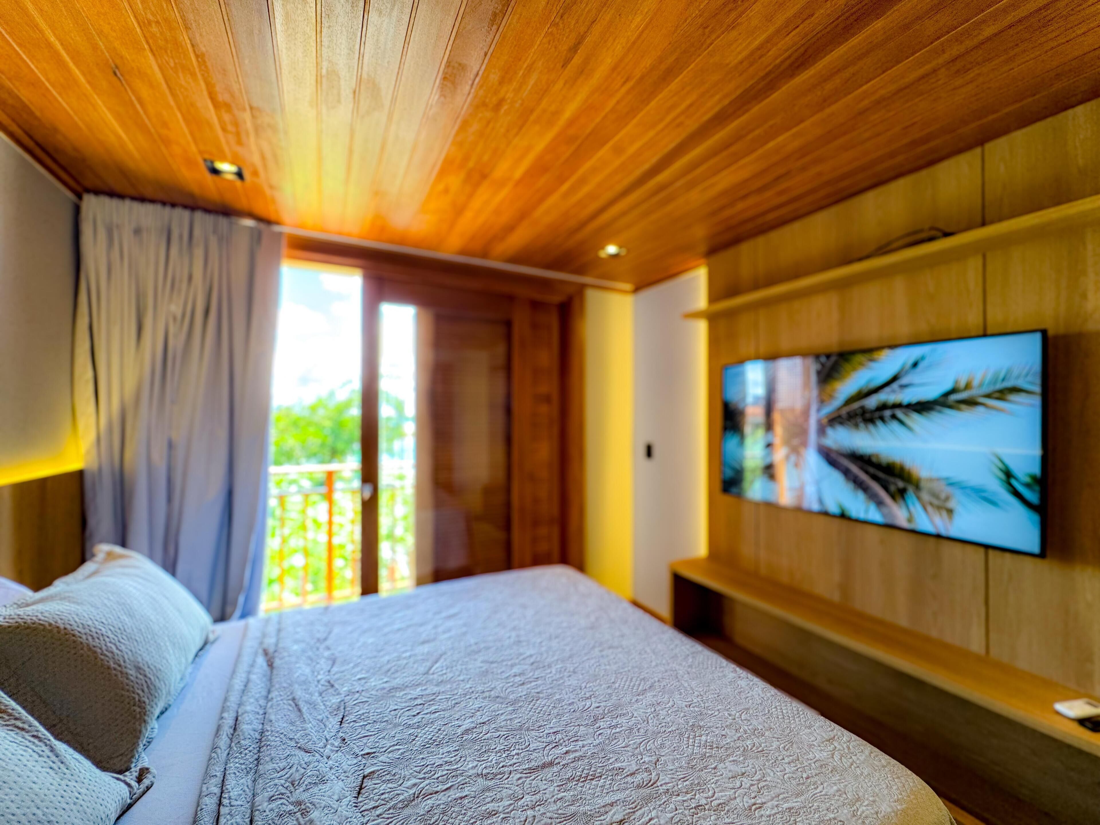 Quarto com cama, televisão fixada na parede e porta de vidro deslizante para uma varanda.