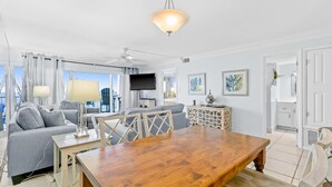 A modern living room with a wooden dining table, grey sofa, and a ceiling fan. - Snowbird-friendly Gulf-front condo with outdoor pool & gym (Miramar Beach)