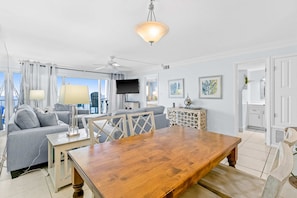 A modern living room with a wooden dining table, grey sofa, and a ceiling fan. - Snowbird-friendly Gulf-front condo with outdoor pool & gym (Miramar Beach)