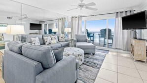 A modern living room with grey sofas, a round ottoman, ceiling fans, a flat-screen TV, and a view of the outdoors. - Snowbird-friendly Gulf-front condo with outdoor pool & gym (Miramar Beach)
