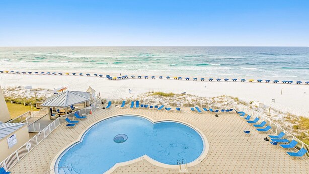 A pool area with a heart-shaped pool, surrounded by blue lounge chairs and umbrellas, overlooking a beach with a sandy shore and ocean waves. - Snowbird-friendly Gulf-front condo with outdoor pool & gym (Miramar Beach)