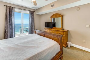 Un dormitorio con una cama grande, un aparador de madera, un televisor y vista al océano a través de un ventanal corredera.