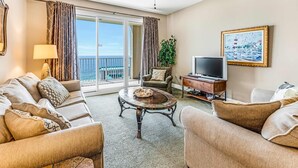 A spacious living room with a sofa set, a coffee table, a TV, and a painting on the wall. - Family-friendly beach condo with coastline views, balcony, shared pool, gym, tennis (Miramar Beach)