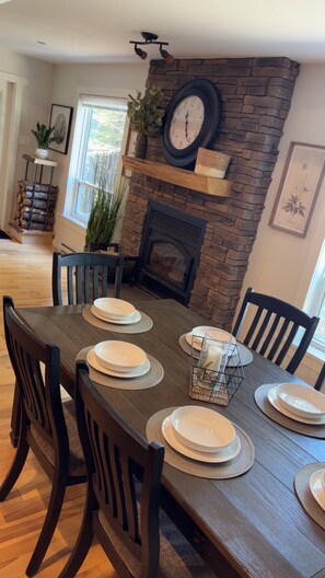 A dining area with a wooden table set for six, surrounded by dark wooden chairs. A stone fireplace with a clock above it is visible. - Waterfront Sunset Cove (Goobies)