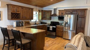 A kitchen with wooden cabinets, a stainless steel refrigerator, a microwave, and a stove. There are two bar stools and a coffee maker on the counter. - Waterfront Sunset Cove (Goobies)