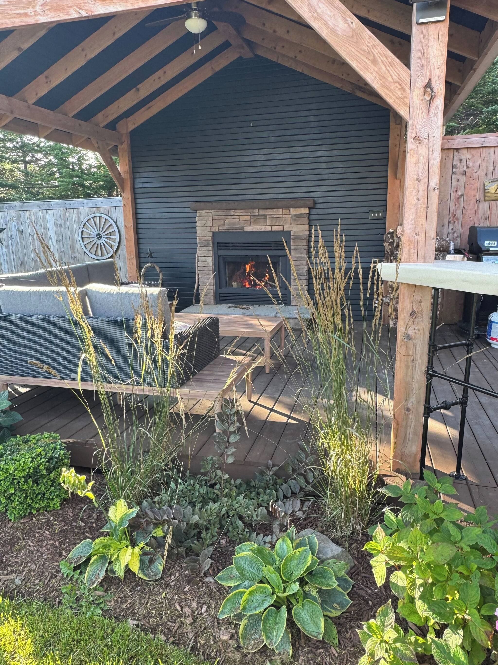 Un espacio al aire libre con techo, que cuenta con una chimenea de piedra, una terraza de madera y diversas plantas.