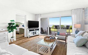 A modern hotel room with a flat-screen TV, a sofa, a dining area with outdoor seating, and a large window offering a view. - Ground floor oceanfront beachy condo with furnished patio, outdoor pool, gym (Miramar Beach)