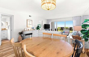 A modern kitchen with a dining area, a television, and a large window offering a view. - Ground floor oceanfront beachy condo with furnished patio, outdoor pool, gym (Miramar Beach)