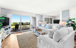 A modern living room with a sofa, television, and a view of the outdoors. - Ground floor oceanfront beachy condo with furnished patio, outdoor pool, gym (Miramar Beach)