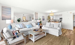 A modern living room with a sofa set, a coffee table, and a dining area with a kitchen in the background. - Ground floor oceanfront beachy condo with furnished patio, outdoor pool, gym (Miramar Beach)
