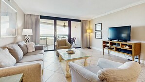 Una habitación de hotel moderna con sofá, sillones, una mesa de centro de vidrio, televisión y balcón con cortinas.