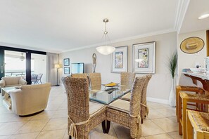 A modern living room with a glass-top dining table surrounded by wicker chairs, a sofa, and a coffee table. There are framed artworks on the walls and a ceiling light fixture. - Beautiful beachfront condo with shared pool and playground (Miramar Beach)