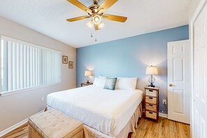 Ein Schlafzimmer mit einem Bett, einem Deckenventilator, einem Nachttisch mit Lampe, einer Tür und einem Fenster mit Jalousien.