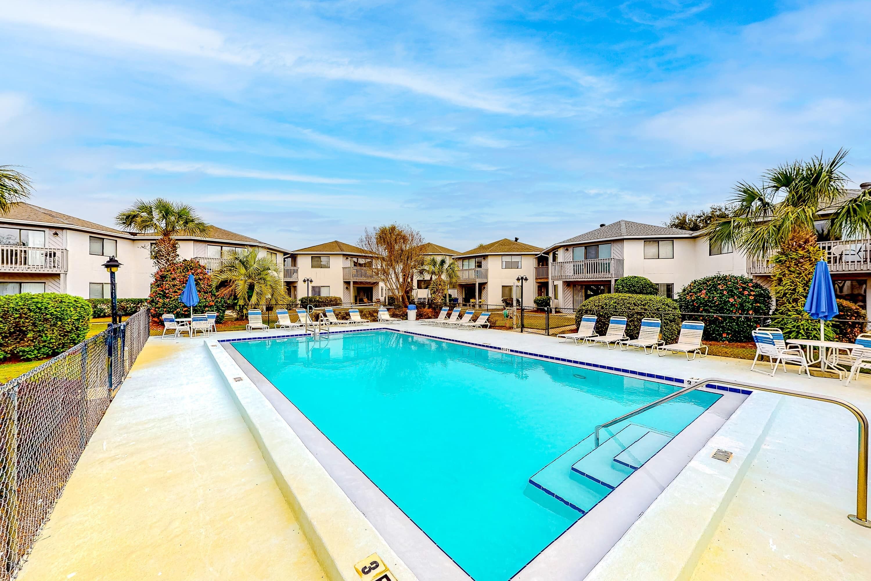 A rectangular swimming pool with a blue interior, surrounded by lounge chairs and umbrellas, with apartment buildings in the background.