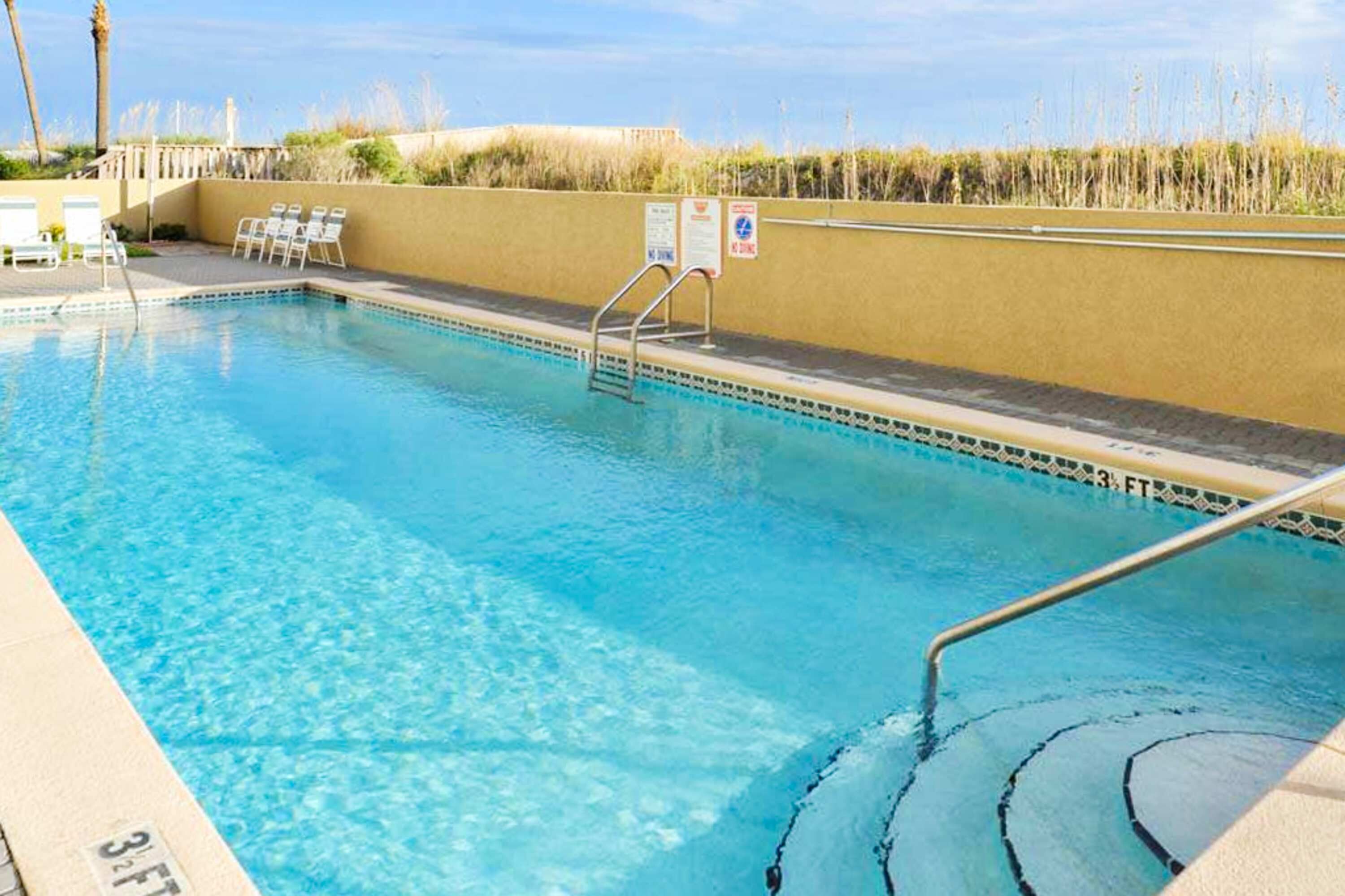 Une piscine d’un bleu éclatant, dont la profondeur maximale est indiquée à 3 pieds, entourée d’un mur de couleur beige et d’une zone herbeuse agrémentée de chaises.