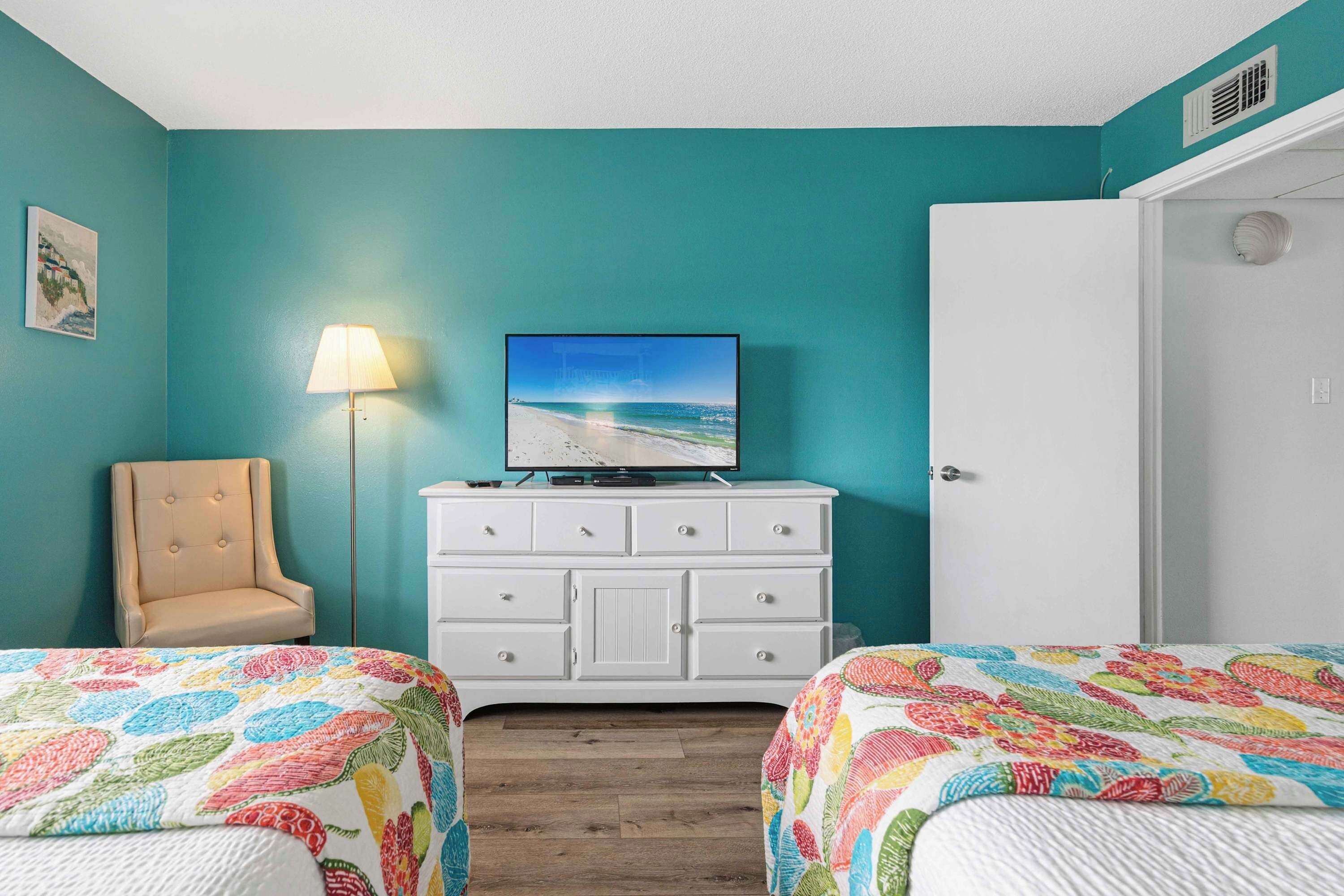 A room with teal walls, a bed with a colorful floral bedspread, a white dresser with a television, a lamp, and a door.