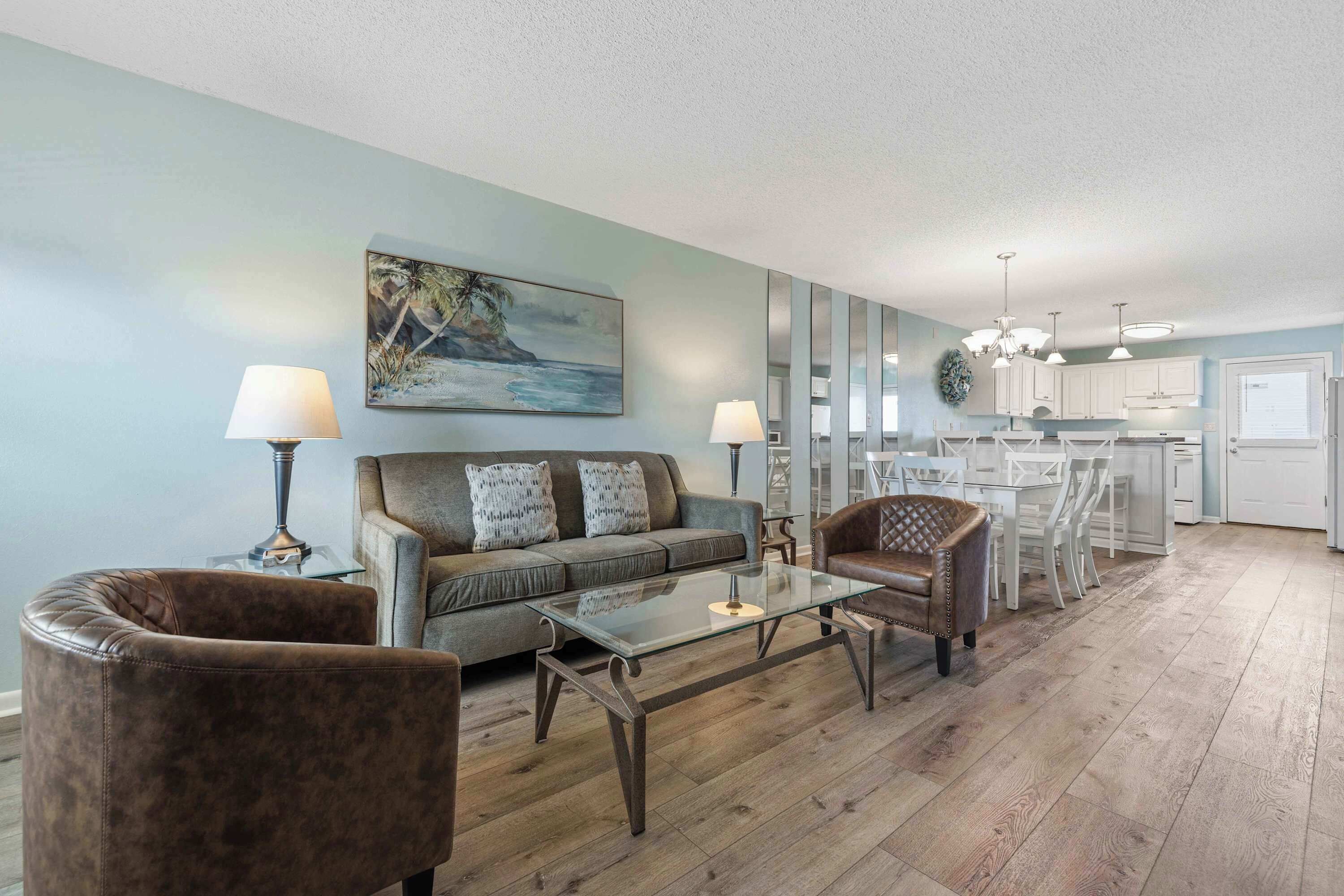 A living room with a sofa, two armchairs, a glass coffee table, and a painting of a beach scene.