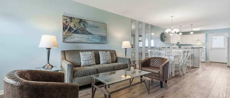 A living room with a sofa, two armchairs, a glass coffee table, and a painting of a beach scene.