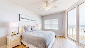 Un dormitorio con una cama grande, ventilador de techo, mesita de noche con lámparas, ventana con persianas y vista a la playa.