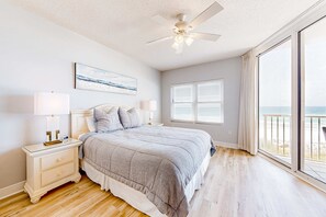 A bedroom with a large bed, a ceiling fan, a nightstand with lamps, a window with blinds, and a view of the beach. - Beachfront condo with balcony, Gulf views, pools & fitness room (Fort Walton Beach)