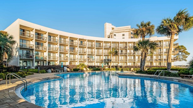 A hotel with a swimming pool surrounded by palm trees and a clear blue sky. - Comfy studio suite with pool & picnic area - snowbird-friendly (Fort Walton Beach)