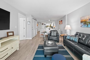 A modern living room with a black leather sofa, a wooden cabinet, and a flat-screen TV. - Sunny, beachfront condo with shared pool, free WiFi, & Gulf views (Fort Walton Beach)