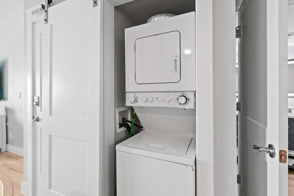 A compact laundry room with a washing machine and dryer, a door with a handle, and a small shelf with a green bottle. - Sunny, beachfront condo with shared pool, free WiFi, & Gulf views (Fort Walton Beach)