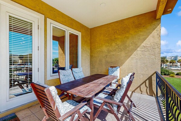 Une terrasse extérieure avec une table et des chaises en bois, de grandes fenêtres équipées de stores et une vue sur les environs.