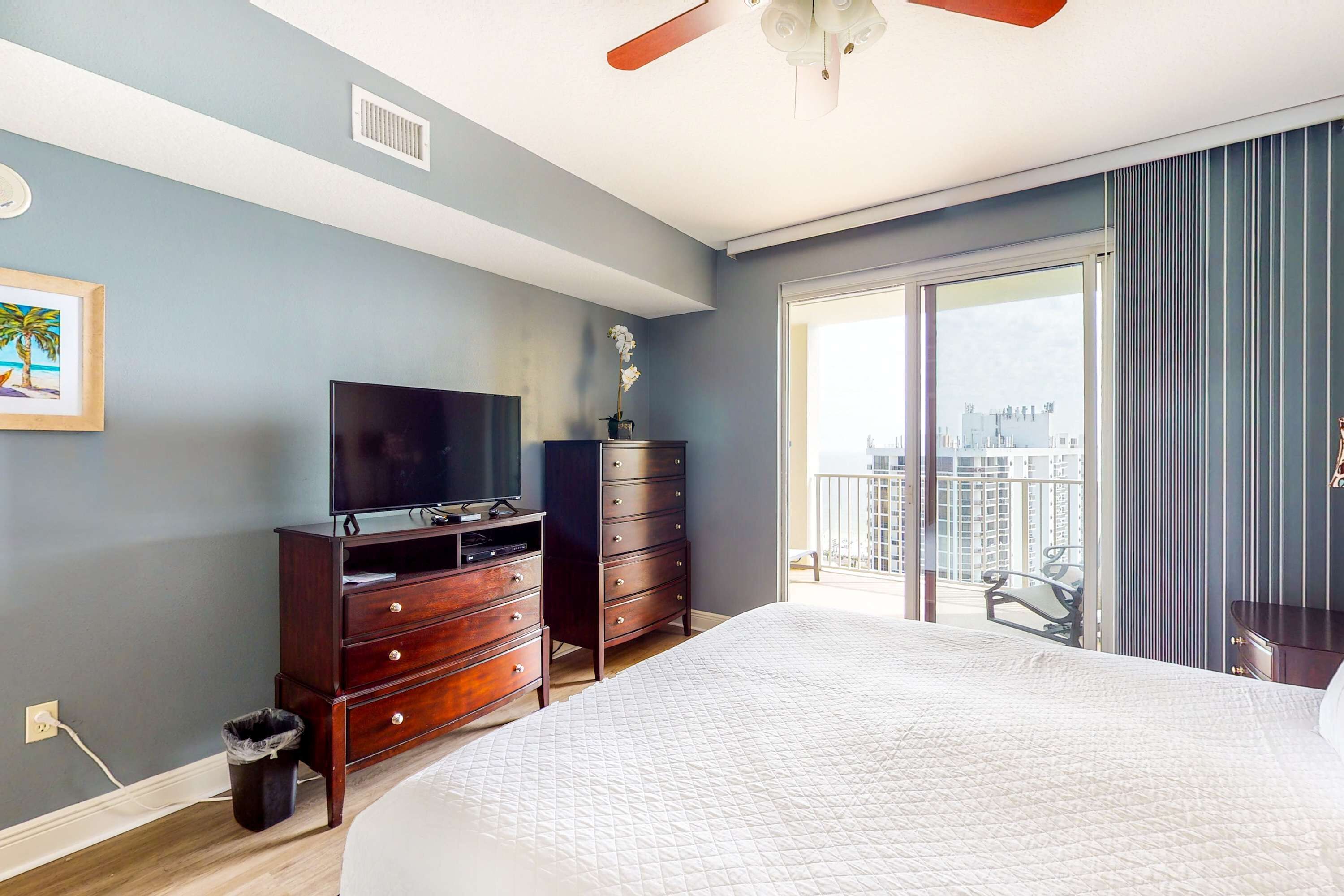 A bedroom with a large bed, a dresser, a television, and a balcony with a city view.