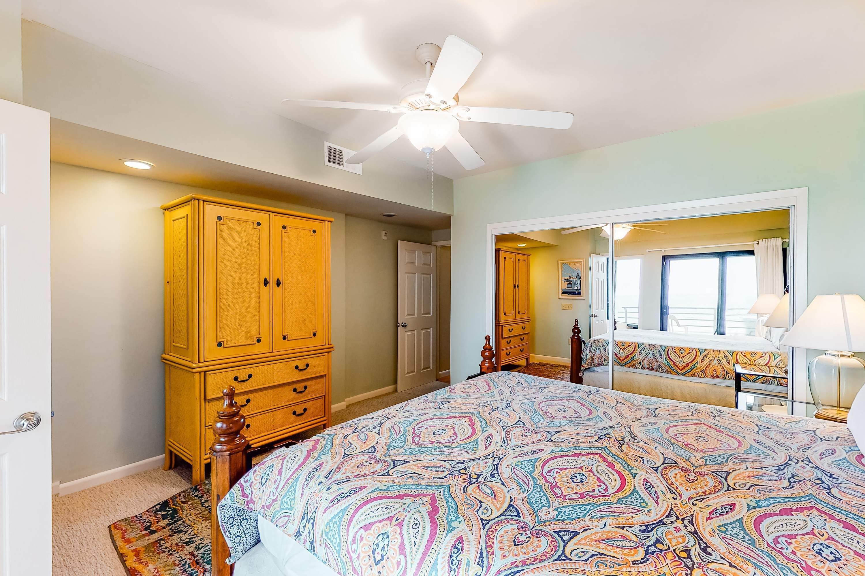 A bedroom with a large bed, a ceiling fan, wooden cabinets, and a view of a balcony.