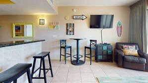 A living area with a bar counter, two bar stools, a round table, two chairs, a sofa with a cushion, a flat-screen TV, and wall decorations including a 'BEACH' sign and a surfboard.