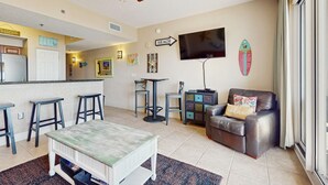 A living room with a TV, a sofa, a dining table, and a kitchen area in the background.