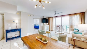 Un vaste espace de vie comprenant une table à manger en bois, des chaises blanches, une console bleue et un ventilateur de plafond.