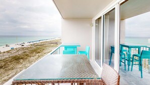 Un balcon avec une table et des chaises en verre, donnant sur une plage où l’on aperçoit des gens et une clôture.