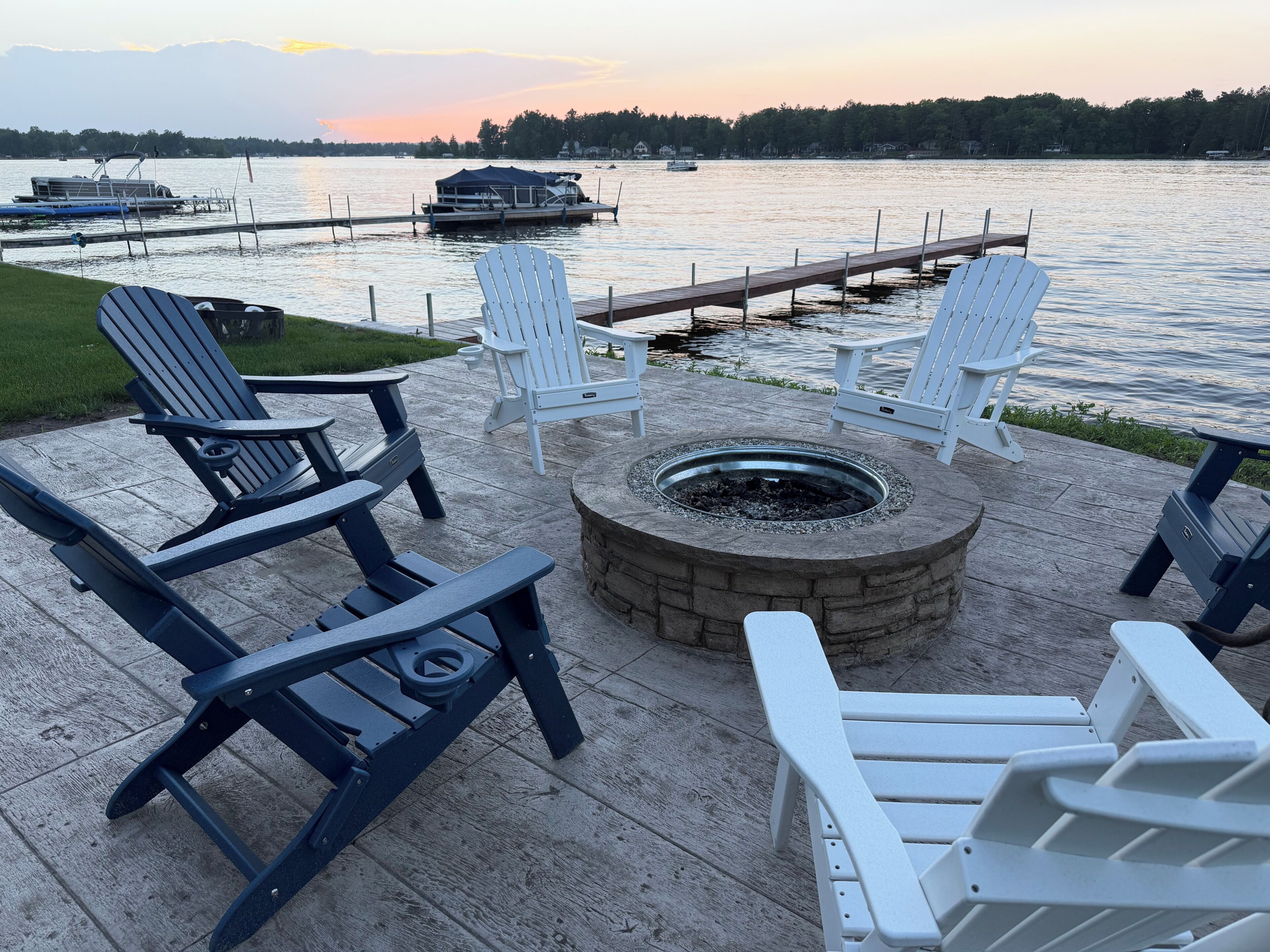A deck with Adirondack chairs, a fire pit, and a view of the lake at sunset.