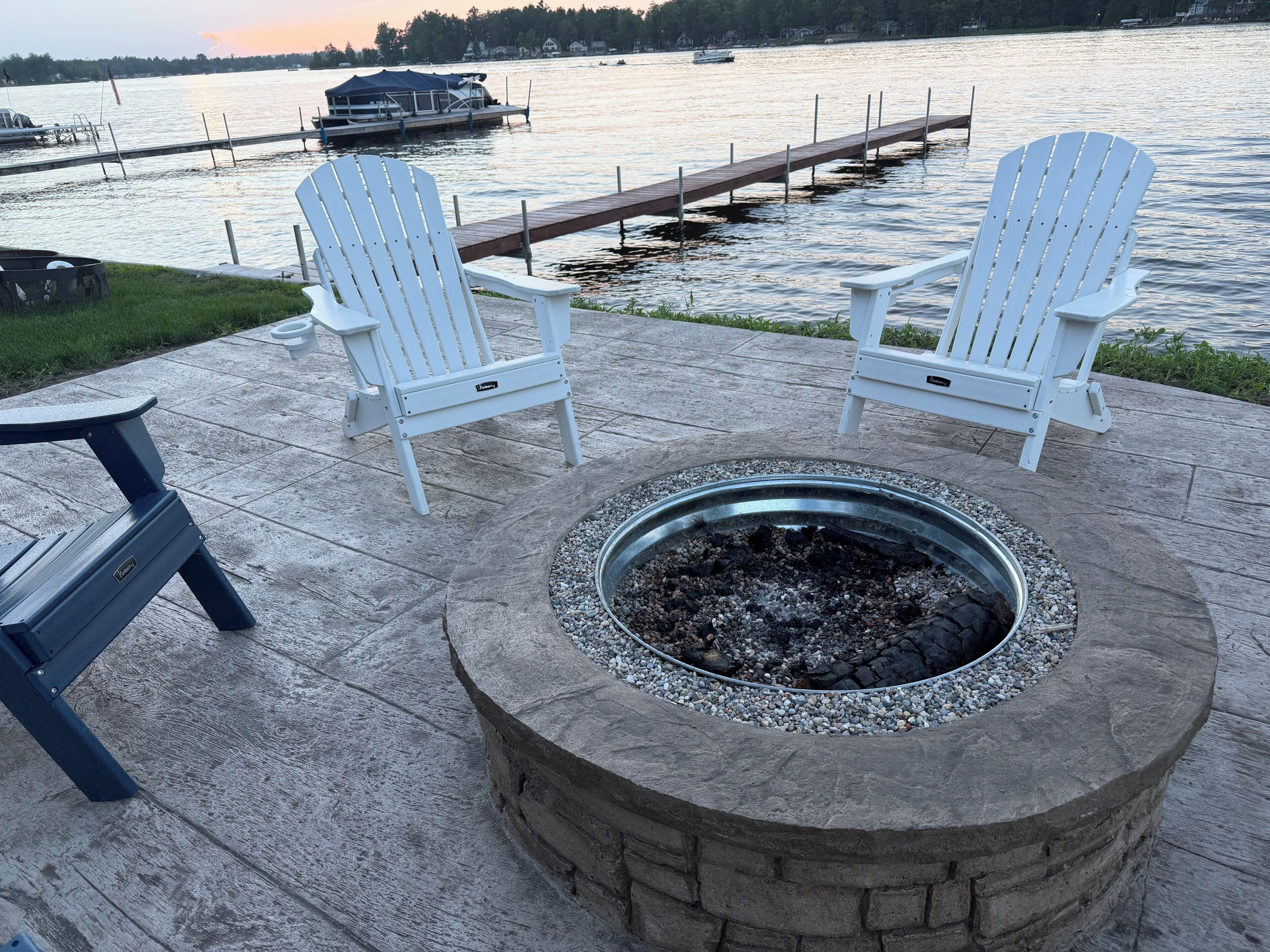 A deck with Adirondack chairs, a fire pit, and a view of a lake with a dock and boats.