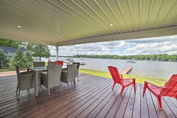 Une terrasse extérieure couverte, aménagée avec des meubles en osier et des chaises rouges, qui offre une vue sur un lac où l’on peut apercevoir des bateaux et un quai.
