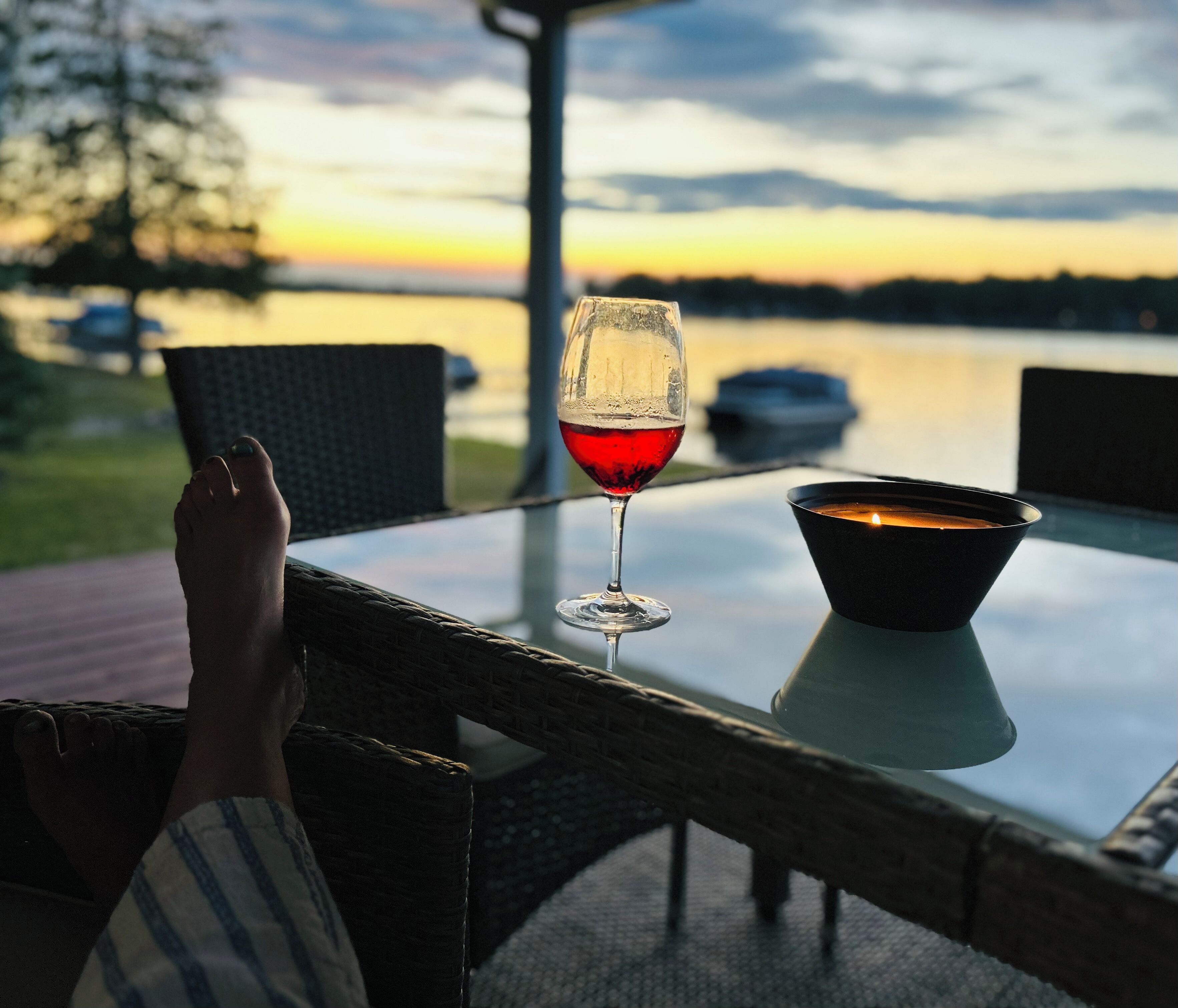 A glass of red wine on a table with a candle, overlooking a body of water and a sunset.