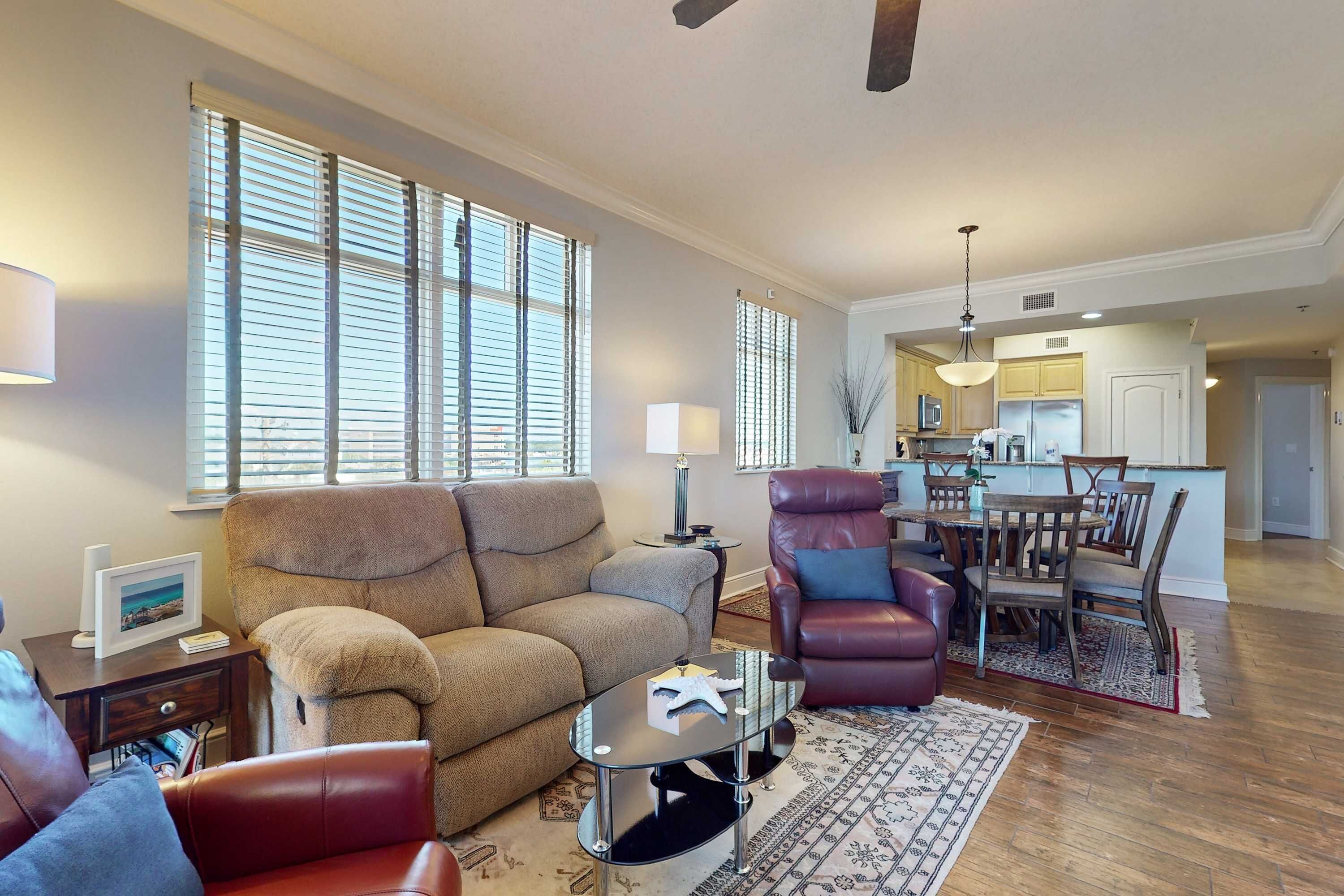 A living room with a sofa, armchair, and coffee table, a dining area with a table and chairs, and a kitchen in the background.
