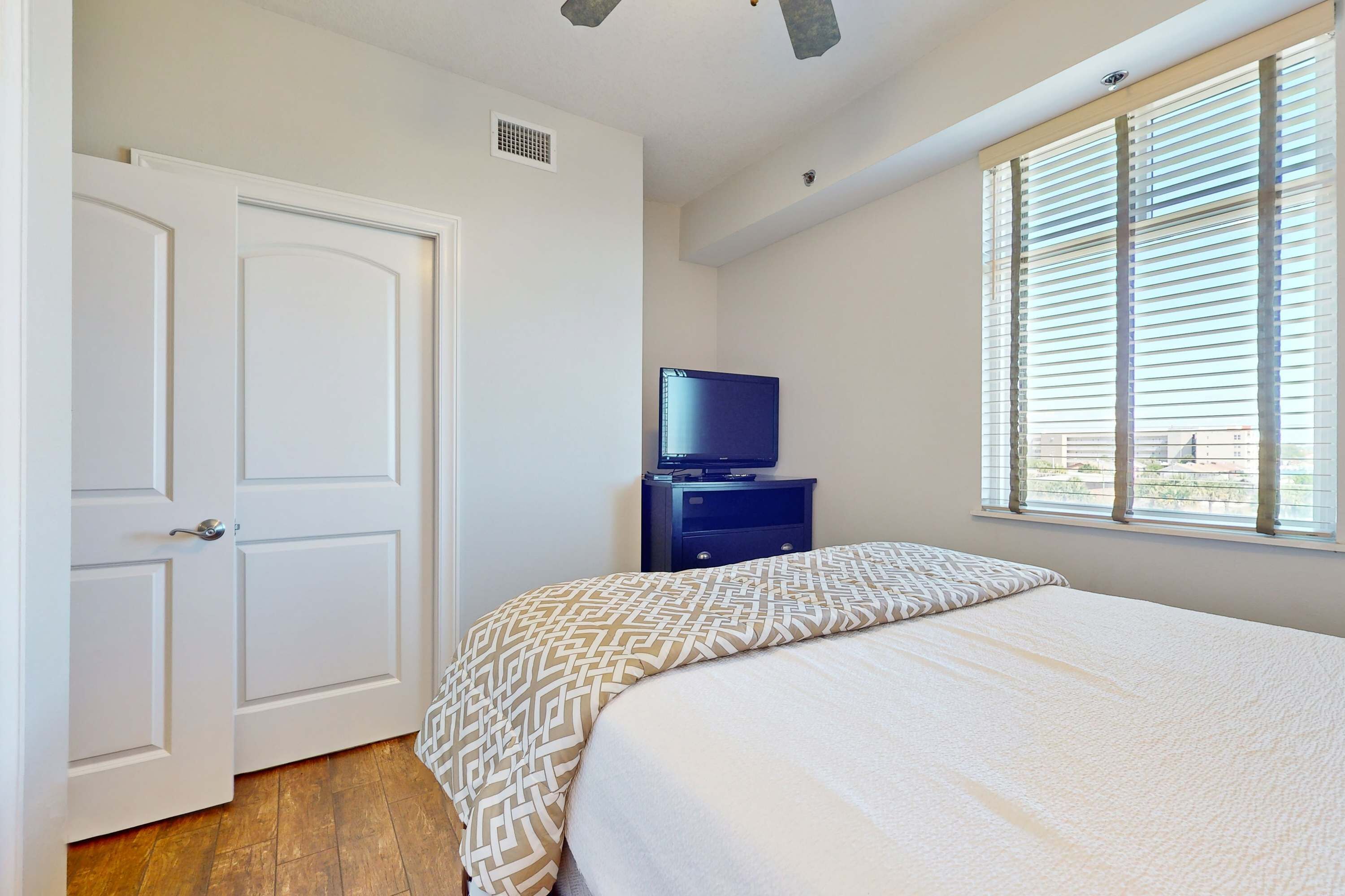 A bedroom with a bed, a television on a stand, a window with blinds, and a door.
