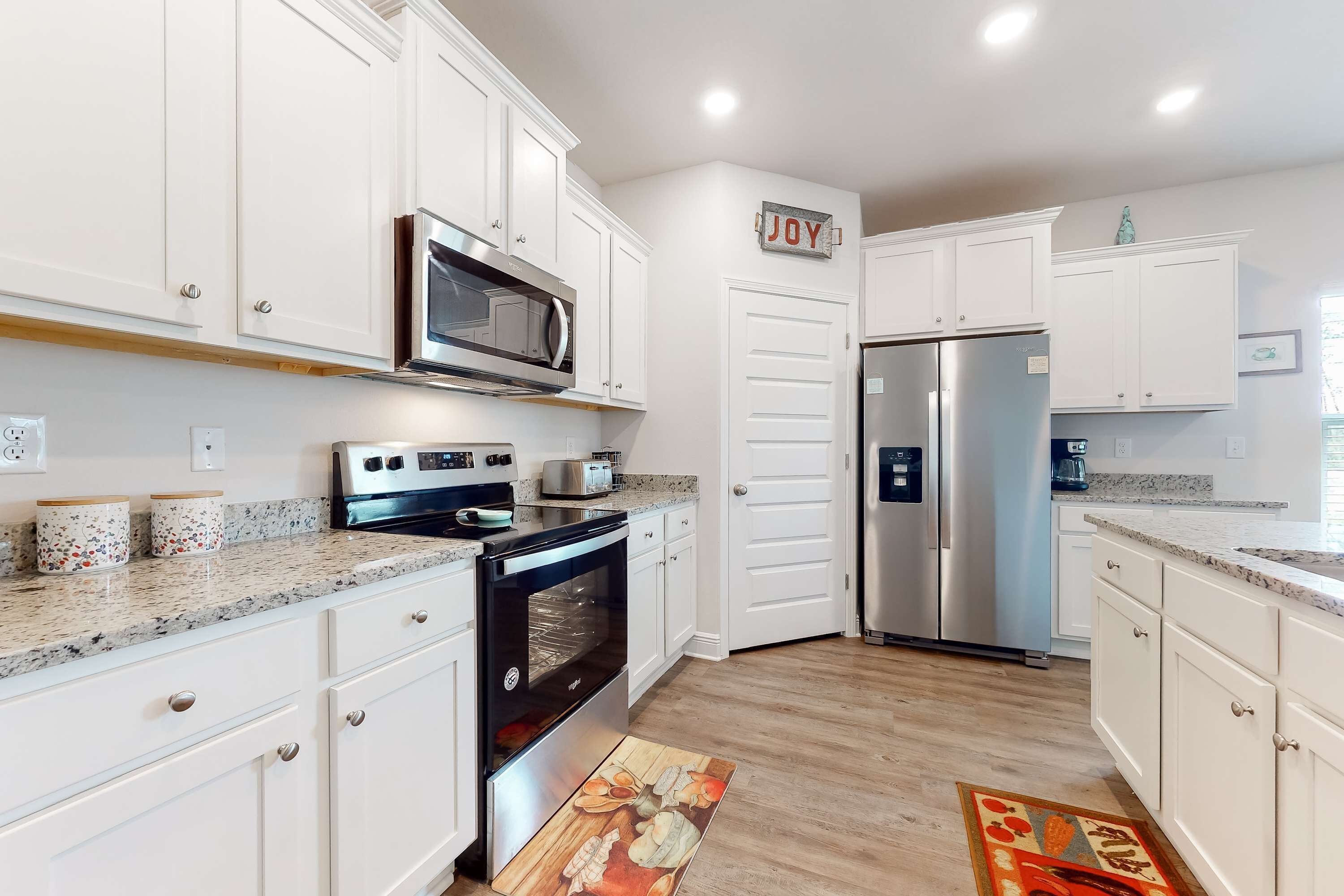 A modern kitchen with white cabinets, stainless steel appliances, and a 'JOY' sign on the wall.